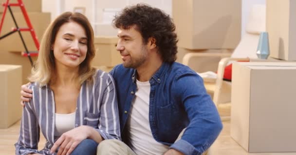 Retrato de la atractiva pareja casada sentados juntos frente a la cámara y sonriendo entre cajas llenas en el nuevo apartamento en un movimiento de día. De interior — Vídeos de Stock