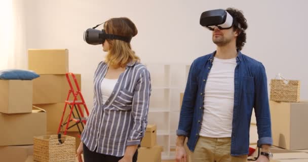 Портретный снимок женатого мужчины и женщины с VR гарнитурой, стоящих в гостиной, полной распакованных коробок в день переезда в новую квартиру. В помещении — стоковое видео
