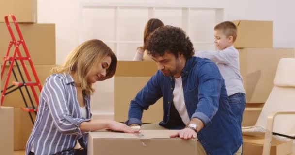 Retrato de la esposa y el marido empacando una caja con una cinta adhesiva y sonriendo a la cámara, luego sus hijos que vienen a ellos desde el fondo. Adentro. — Vídeos de Stock