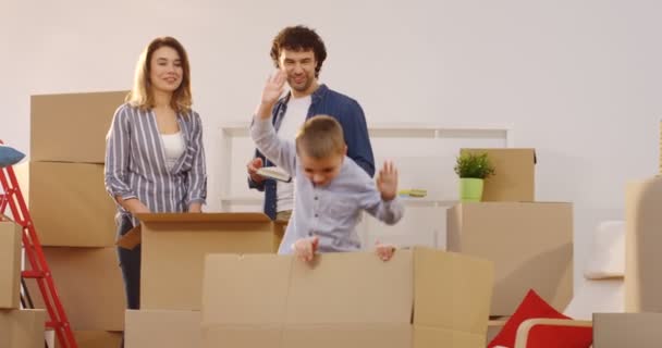 Glada föräldrar står ett trevligt vardagsrum med full av rutorna uppackad med hem grejer när barnen hoppar ur lådan. Inflyttning i nya huset. Inomhus — Stockvideo