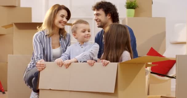 Glückliche, gut aussehende Eltern, die mit ihren Kindern zwischen Kisten sitzen, lächeln und den Daumen nach oben zeigen. Kinder, die in der Box sitzen. Tageseinzug. innen — Stockvideo