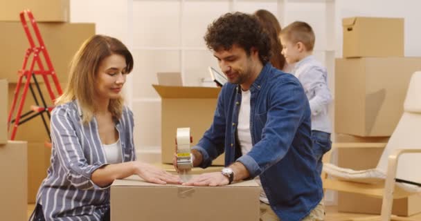 Retrato de la pareja casada empacando una caja con una cinta adhesiva y sus hijos viniendo a ellos desde el fondo para posar juntos frente a la cámara. En interiores — Vídeos de Stock