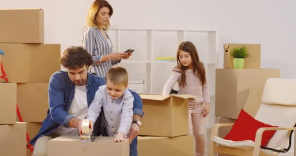 Bra ser far med en liten son som packa en låda med en tejp och en mamma med en dotter på bakgrunden. Flytta ut huset. Inomhus — Stockvideo