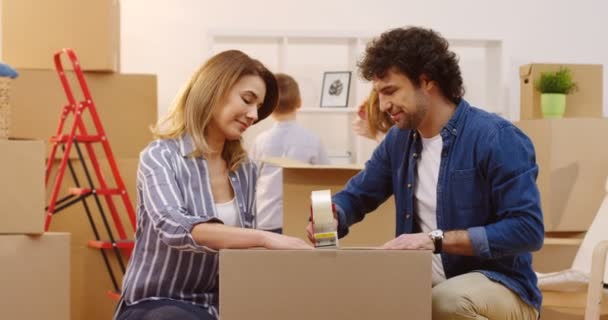 Retrato filmado do homem e da mulher casados embalando uma caixa com uma fita adesiva e conversando com seus filhos no fundo. Interior — Vídeo de Stock