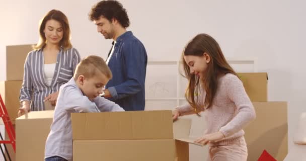 Irmã e irmão bonito desempacotar uma caixa como eles se movendo no novo apartamento, enquanto sua mãe e pai fazendo o mesmo no fundo. Interior — Vídeo de Stock