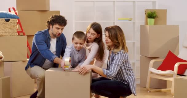 Портрет счастливой семьи родителей и двух детей, упаковывающих коробку с липкой лентой при выезде из квартиры. Внутри — стоковое видео