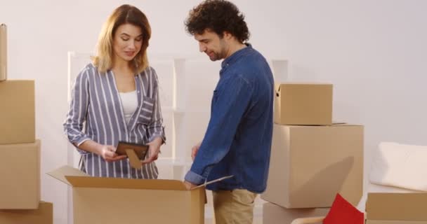 Attractive married man and woman unpacking boxes with different home stuff in their new flat while moving in it. Talking and laughing. Inside — Stock Video