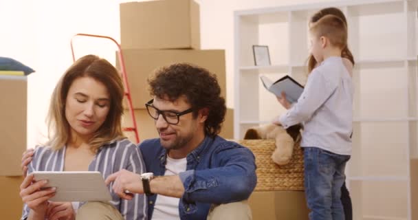 Tablet bilgisayar seyir ve konuşmaya çift Beyaz aile portre çekim onların oğulları ve kızları yeni dairede taşıma sırasında kutuları açma tamamlayın. Kapalı — Stok video