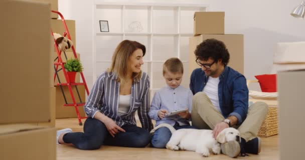 Smiled parents sitting on the floor with a cute little son, talking and watching him playing on the tablet computer. Labrador dog sleeping beside. Portrait shot of the family surrounded by boxes — Stock Video