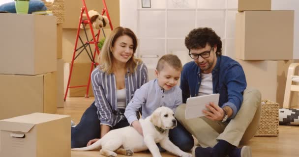 Madre felice, padre e figlio seduti sul pavimento con il loro cucciolo tra scatole di cartone e avendo videochat, agitando le mani e parlando. Interni — Video Stock