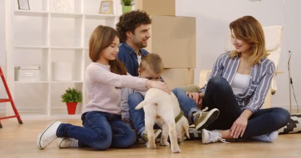 Portraitaufnahme der glücklichen kaukasischen Familie mit einem Labrador-Welpen, der auf dem Boden sitzt, umgeben von den vielen Kisten und Hausrat während des Umzugs in die neuen Häuser. Innenräume — Stockvideo