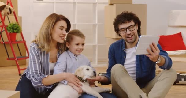 开朗的父母与可爱的儿子和小拉布拉多坐在地板之间的纸箱与家庭的东西和 videochatting, 而移动在新的公寓。肖像拍摄。内 — 图库视频影像