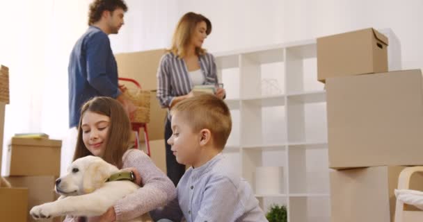 Süße kleine Geschwister, die mit einem Welpen vor der Kamera spielen, während ihre Eltern hinter ihnen Sachen auspacken. Einzug. Innenräume — Stockvideo