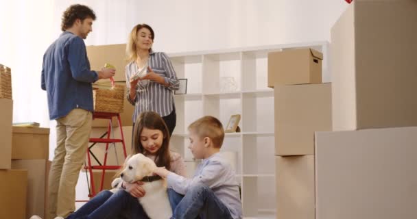 Šťastný malý chlapec a dívka si hraje s štěně labradora na podlaze, zatímco jejich rodiče vybalování krabic na pozadí. Přesouvání in nový dům. Uvnitř — Stock video