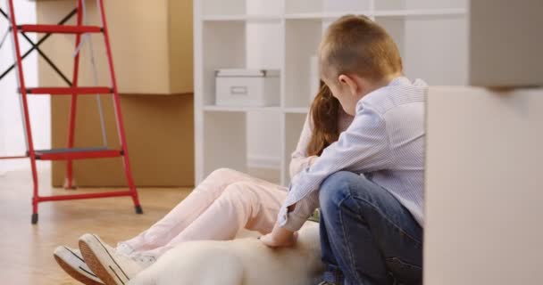 Крупним планом мила дівчина і хлопчик грають з цуценям-лабрадором серед картонних коробок з домашніми речами у вітальні нової квартири. Всередині — стокове відео