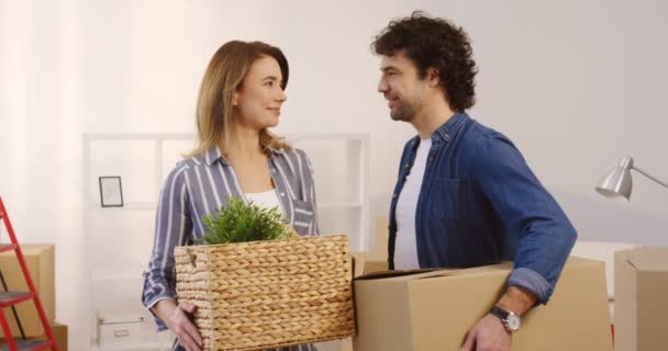 Retrato da mulher de meia-idade casada e homem olhando um para o outro e, em seguida, sorrindo para a câmera enquanto se move no novo apartamento e segurando planta e caixa nas mãos. Lá dentro. Fechar . — Vídeo de Stock