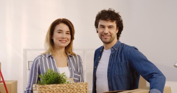 Casal bonito olhando um para o outro e, em seguida, para a câmera enquanto se move no novo apartamento e segurando planta e caixa nas mãos. Dentro de casa. Retrato. de perto . — Vídeo de Stock