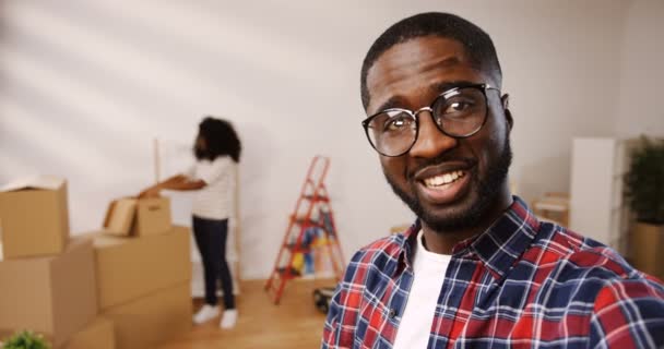 Dekat dengan pemuda Afrika Amerika berkacamata yang sedang merekam video sambil pacarnya membongkar kotak saat mereka pindah ke apartemen baru. Kemudian dia datang ke kamera, memeluknya dan mereka — Stok Video
