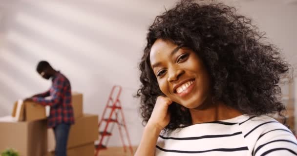 Κοντινό πλάνο του σγουρά νεαρός αφρικανική αμερικανική γυναίκα έχοντας videochat και το φίλο της σε ποτήρια αποσυσκευασία κιβωτίων πίσω κατά τη διάρκεια μετακίνηση στο νέο σπίτι τους. Τότε ο άνθρωπος έρχεται σε αυτήν και να μιλάμε με χαμόγελο — Αρχείο Βίντεο