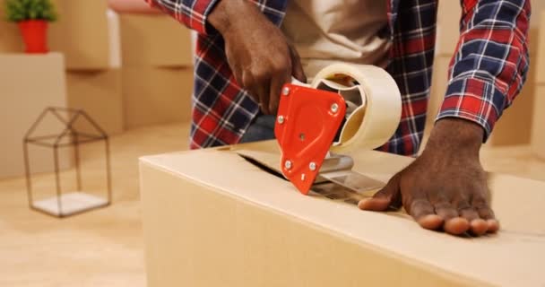 Primer plano de las manos masculinas afroamericanas empacando una caja con una cinta adhesiva en la habitación llena de cajas. Adentro. — Vídeo de stock