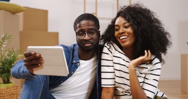 Portrait de jeune couple afro-américain assis sur le sol et vidéoconférence sur la tablette entre les boîtes tout en se déplaçant dans la nouvelle maison. Intérieur — Video