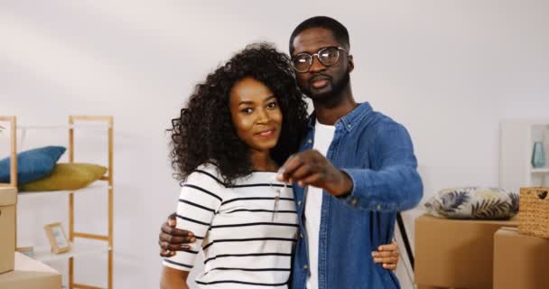 Porträtt av den glada unga afroamerikanska gifta bara par kramar framför kameran och man visar nycklar från deras hem i handen. Flytta i nya lägenheten. Inomhus — Stockvideo
