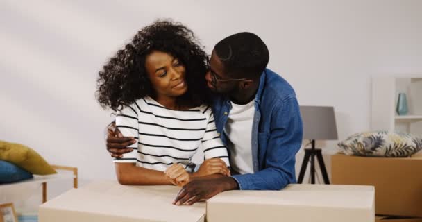 Retrato de la feliz sonrisa afroamericana recién casada pareja apoyada en las cajas de la sala de estar, mirándose y abrazándose delante de la cámara mientras se mueven en su casa. En interiores — Vídeo de stock