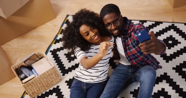 Vista de cima sobre o feliz jovem afro-americano recém-casado casal sentado no chão coberto com tapete estampado perto de uma caixa e fazer selfies com as chaves nas mãos no smartphone. Interior — Vídeo de Stock