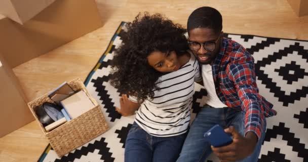 Vista desde arriba de los jóvenes y alegres afroamericanos recién casados hombre y mujer sentados en el suelo cubiertos con alfombras estampadas y haciendo buenos selfies en el teléfono inteligente. Adentro. — Vídeo de stock