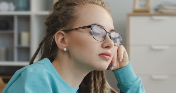 Närbild av den unga vackra kvinnan med dreadlocks i glasögon och blå skjorta ser till kameran i mysiga rum. Porträtt. Inomhus — Stockvideo