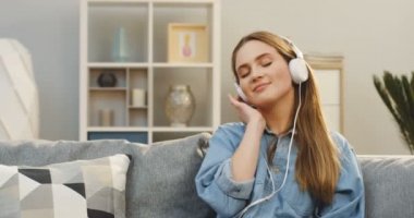 Beyaz kulaklık Smartphone müzik dinlemek ve evde kanepede otururken gülümseyen sevimli genç kadın portresi. Kapalı