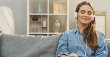 Kot gömlek ve beyaz kulaklık rahat daire içinde kanepede otururken Kapalı gözlerle onun smartphone üzerinde müzik dinlemekten zevk gülümsüyor genç kadın portre çekim