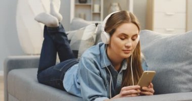 Beyaz kulaklık müzik onun smartphone üzerinde rahat oturma odasında kanepede yatan dinlemekten zevk genç ve güzel kadın portresi. Kapalı