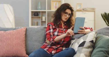 Ekose gömlek ve rahat oturma odasında kanepede otururken tablet cihaz üzerinde kayıt gözlük güzel ve genç birbayan. Kapalı