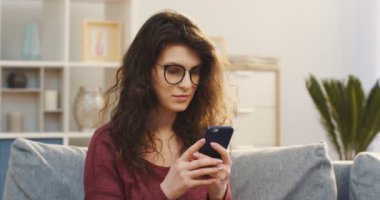 O zaman evde kanepede otururken düşünüyorum çekici genç kıvırcık kadın bardaklarda kaydediyor ve onun smartphone üzerinde kaydırma portresi. Kapalı