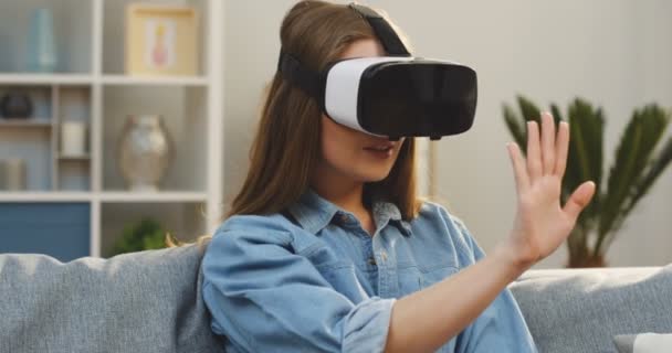 Портрет молодої жінки, яка сидить на дивані і має гарнітуру VR вдома. VR окуляри. Внутрішні — стокове відео