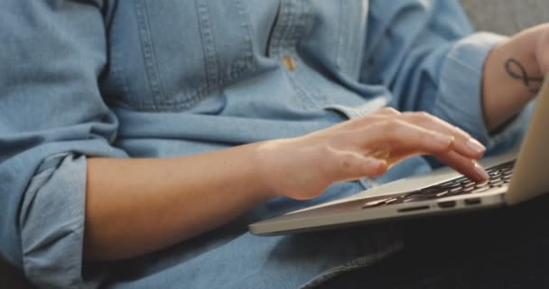 Primer plano de las manos femeninas con camisa vaquera trabajando y grabando en la computadora portátil. En casa, en el sofá. En interiores — Vídeos de Stock