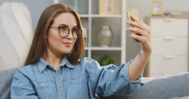 Primer plano de la joven mujer bonita en las gafas haciendo una selfie en el teléfono inteligente en el sofá en la acogedora habitación. Adentro. — Vídeos de Stock