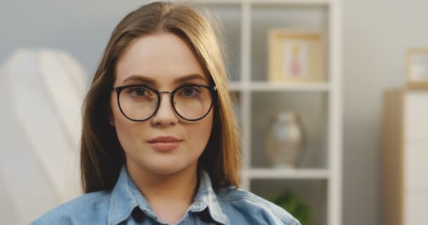 Retrato de la joven y encantadora mujer con gafas mirando a la cámara y riendo alegremente en la sala de estar. De cerca. Adentro. — Vídeo de stock