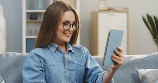 Retrato de la joven atractiva en la camisa jeans y gafas que tienen video chat en la tableta de la acogedora sala de estar. De interior — Vídeos de Stock
