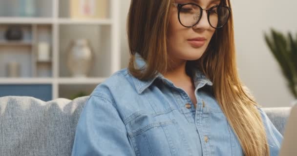 Retrato filmado da jovem bonita na camisa jeans e óculos trabalhando no computador portátil em casa e, em seguida, girando a cabeça com um sorriso para a câmera. Fechar — Vídeo de Stock