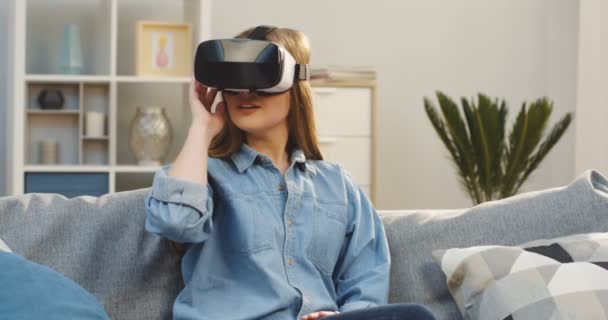 Ung kvinna i jeans skjorta och Vr-glasögon sitter i soffan och ha ett Vr-headset i vardagsrummet. Inomhus — Stockvideo