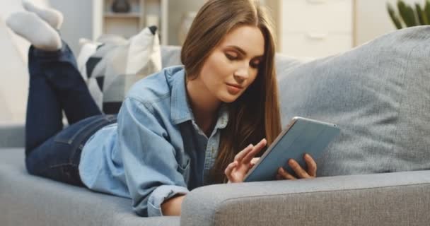 Schöne junge Frau blättert auf dem Tablet-Computer und lächelt, während sie zu Hause auf dem Sofa liegt. innen — Stockvideo