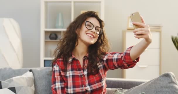 肖像的年轻卷曲的妇女在眼镜和格子衬衫做一个自拍的智能手机上的沙发上, 在舒适的房间。室内 — 图库视频影像