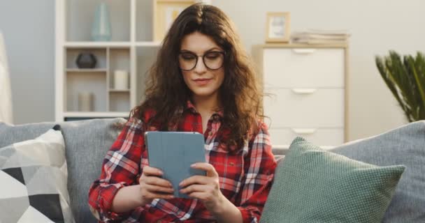 Aantrekkelijke jonge vrouw in de bonte shirt en glazen scrollen op de tablet pc en surfen op het Internet in de gezellige woonkamer. Indoor — Stockvideo
