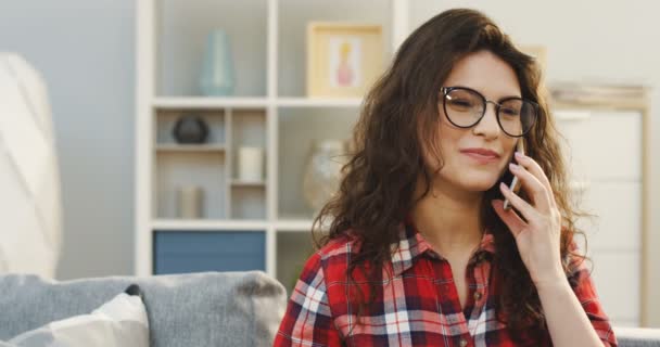 Gros plan de la jeune femme frisée belle dans les lunettes et chemise bigarrée parler sur le smartphone dans le salon confortable. À l'intérieur. Portrait — Video