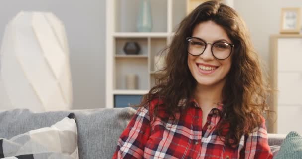 Közelkép a bájos fiatal nő szemüveg és kockás ing néz egyenesen a kamerába, és mosolyogva a lakás szoba. Portré. Belső — Stock videók