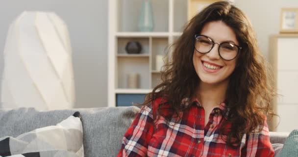 Portret van de jonge mooie vrouw in glazen en plaid shirt op zoek naar de camera en vreugdevol lachen in de gezellige kamer. Close-up. Binnenkant — Stockvideo