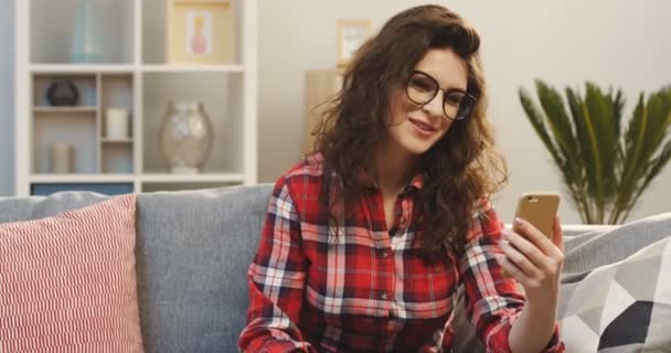 格子縞のシャツと居心地の良いリビング ルームでスマート フォンでビデオ チャットを持つメガネの魅力的の若い女性の肖像画。屋内 — ストック動画