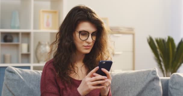 Πορτρέτο της η ελκυστική νεαρή γυναίκα σγουρά στα γυαλιά taping και κύλιση στο smartphone της, τότε σκέψης ενώ κάθεται στον καναπέ στο σπίτι. Σε εσωτερικούς χώρους — Αρχείο Βίντεο